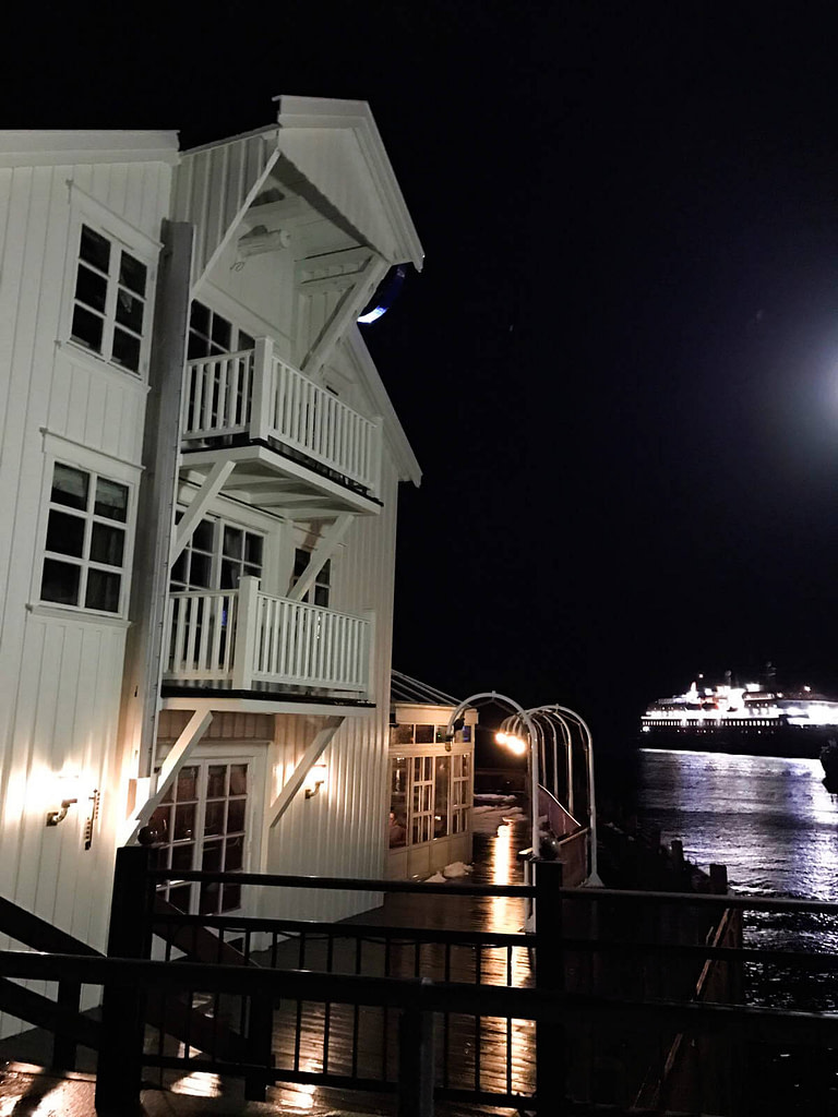 Retro di un edificio bianco in legno e vista sull'Hirtigruten illuminato attraccato al porto di Svolvær